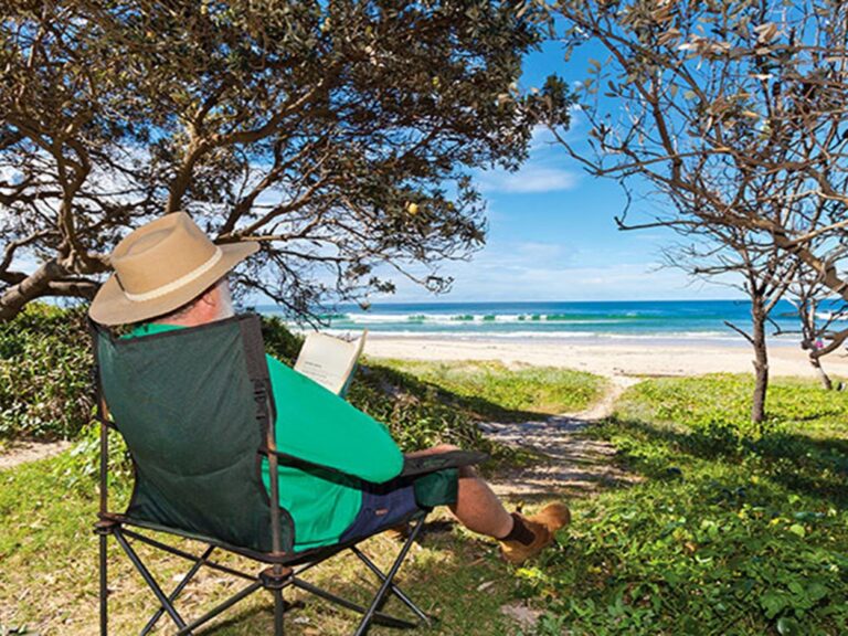 A man reads a book in the shade with a view of the beach in Red Cliff campground, Yuraygir National